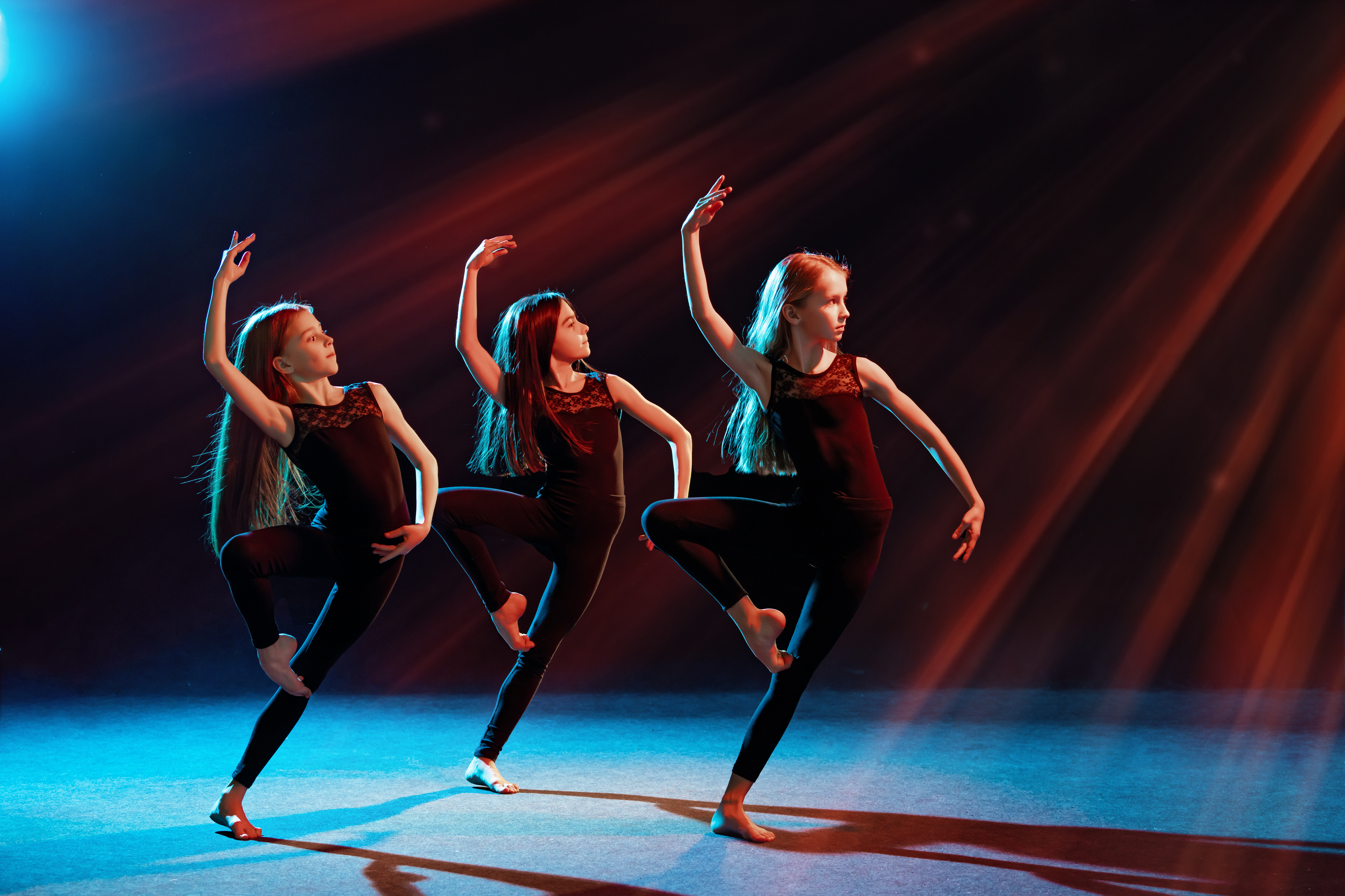 group of three ballet girls in tight-fitting costumes dance against black background with their long hair down, silhouettes illuminated by color 