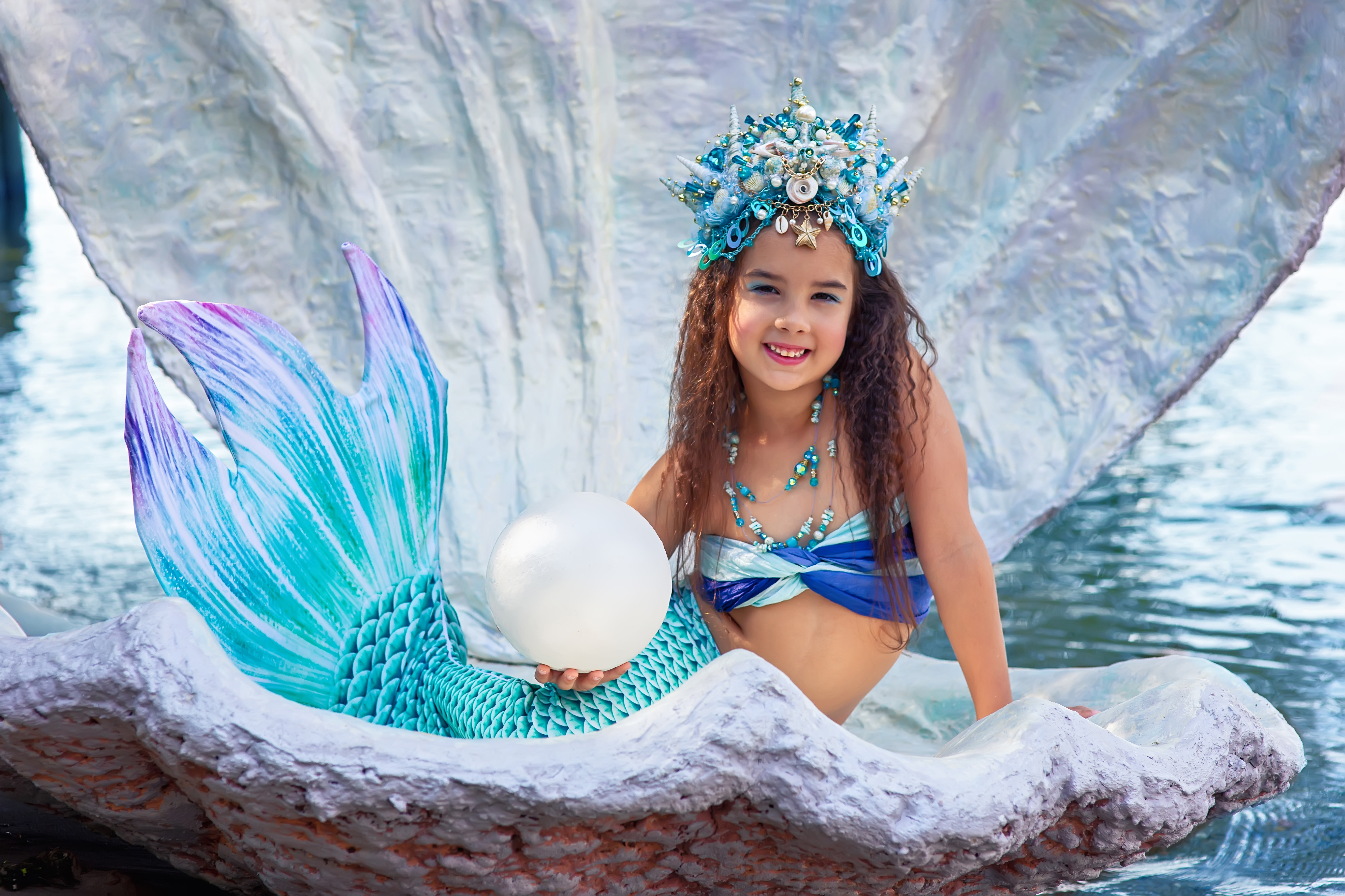 A beautiful happy little girl in a mermaid costume sits in a large sea shell